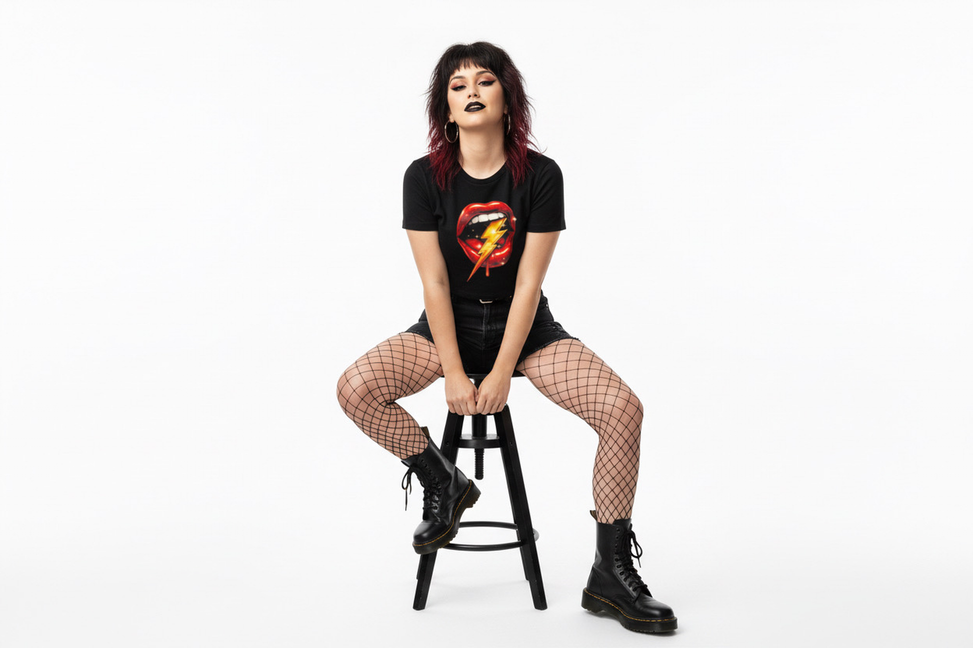 Person sitting on a stool wearing a black t-shirt with a graphic design, fishnet stockings, and black boots on a white background.
